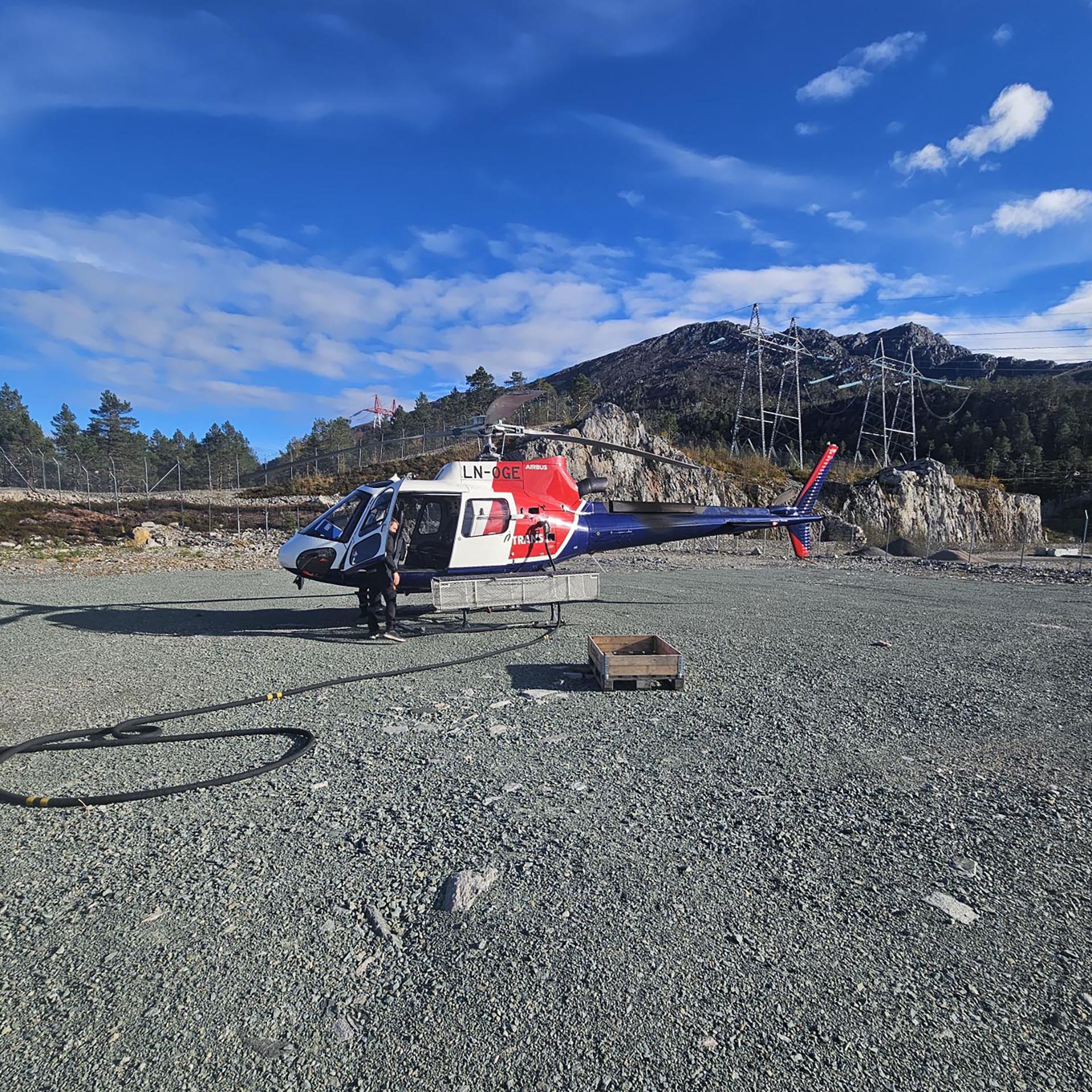 Helikopter som står parkert på en stor grusplass med synlige høyspentlinjer i bakgrunnen. Døra til helikopteret er åpent, og foran står det en mann som er på vei inn.