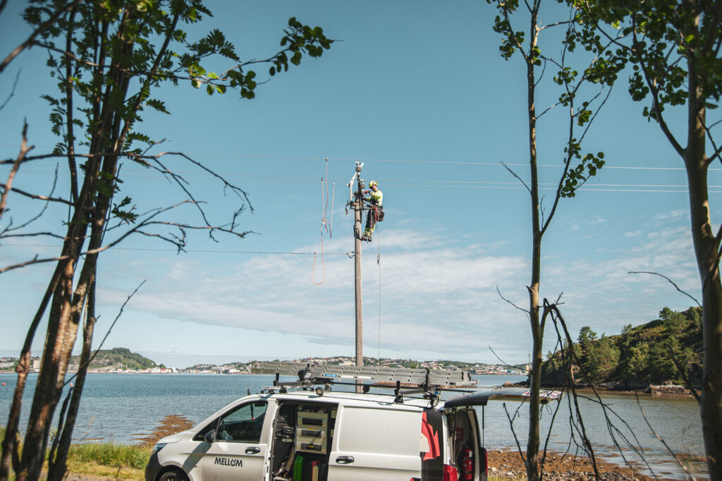 Energimontør øverst i ei mast med nydelig vær i bakgrunnen og en Mellom arbeidsbil i front.